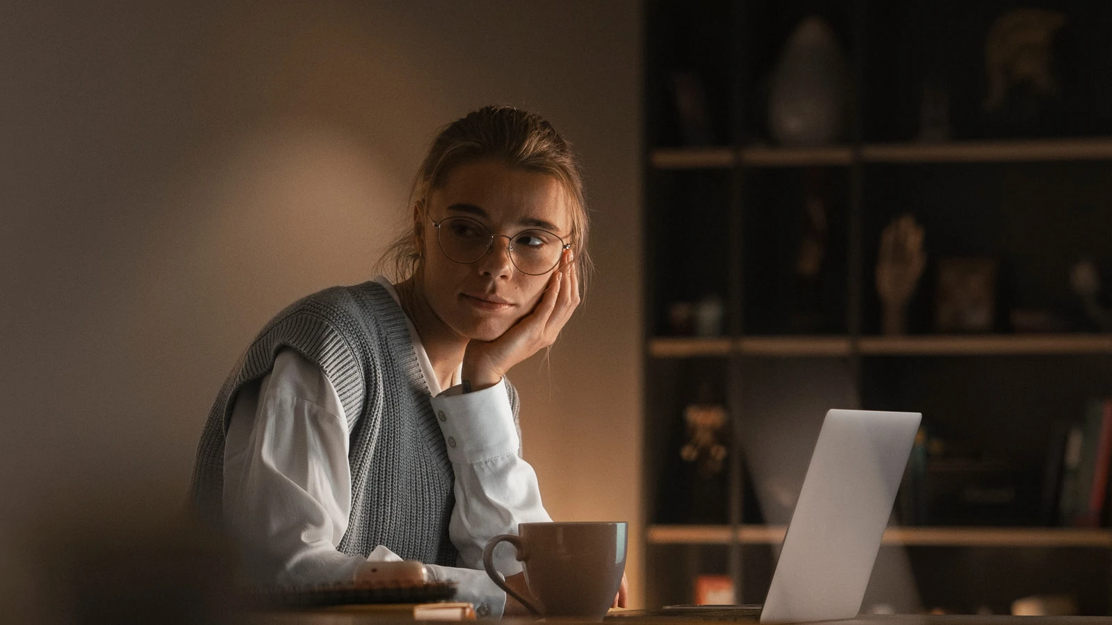 Mulher de óculos apoia o rosto na mão diante de um notebook, com uma xícara na mesa, numa sala iluminada por um abajur à noite